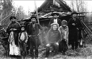 Seven indigenous people ranging in age from children to very elderly sit and stand in front of a rustic shack.